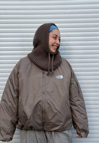 Brun polstret jakke med høj krave og lynlås, med The North Face-logo. Personen bærer en strikket brun hætte og grå bukser.