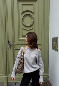 Pull gris à épaules dénudées, pantalon noir ajusté, portant un sac à main marron à motifs ; porte verte ornée en arrière-plan avec un interphone mural.