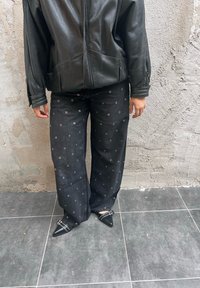 Schwarze Lederjacke, locker sitzende schwarze Denim-Hose mit silbernem Ösenmuster und spitze schwarze Schuhe mit Verzierungen.