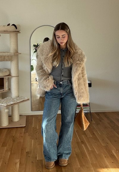 Abrigo de piel sintética beige, cárdigan gris, jeans acampanados de color azul claro y botas beige; sosteniendo un bolso marrón, con un espejo y una planta de fondo.