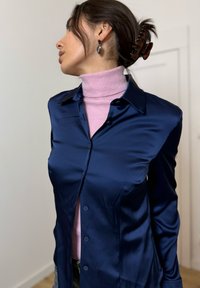 Temno modra satenasta srajca z gumbi in ovratnikom, pod njo je zataknjen roza rebrasti pulover z visokim ovratnikom; oblečena s sivo hlačo. Barvni kontrast je opazen.