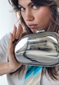 Fémes ezüst clutch táska lekerekített formával és sima felülettel. Kicsi felső csattal rendelkezik, és derékszintén tartják egy világos háttér előtt.