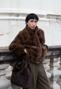 Person med mørkt krøllet hår læner sig på en stensbalustrade, if�ørt en brun pelsfrakke og holder en matchende pels-håndtaske udendørs.