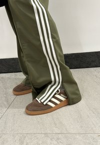 Ελαιόλαδο πράσινα αθλητικά παντελόνια με λευκές ρίγες και πλαϊνούς φερμουάρ, συνδυασμένα με καφέ σουέτ αθλητικά παπούτσια που έχουν λευκές λεπτομέρειες και λογότυπο.