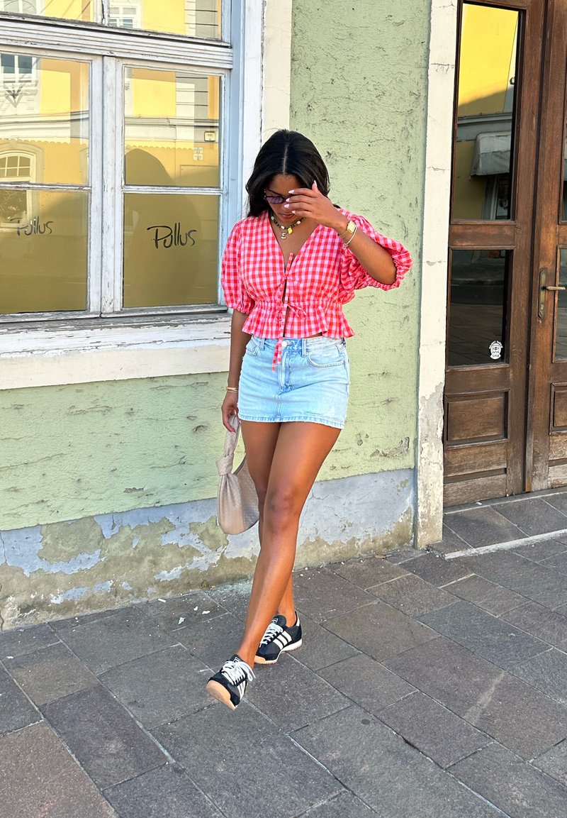 Cropped blouse in red and white gingham with puff sleeves, teamed with light denim shorts. The outfit is complemented by black sneakers and a beige handbag.