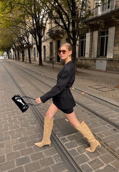 Blazer negro ajustado sobre un vestido mini negro, combinado con botas de ante beige hasta la rodilla. Se sostiene un pequeño bolso negro en la mano.
