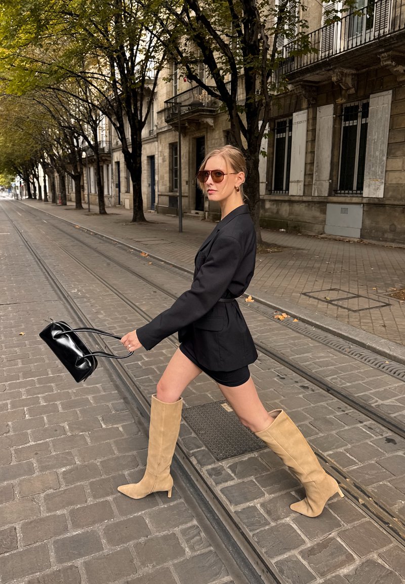 Black tailored blazer over a black mini dress, paired with knee-high beige suede boots. A small black handbag is held in the hand.