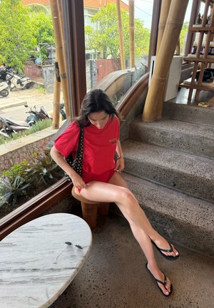 Joven mujer con camiseta roja y pantalones cortos sentada en un taburete de madera junto a grandes ventanas, ajustándose los pantalones cortos con unas gafas de sol sobre una mesa cercana.