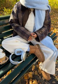 Rjava karirasta jakna na gumbe preko bele pletene majice, kombinirana s kremnimi širokimi hlačami in rjavimi semiš čevlji. Modra ruta in slušalke v bližini.