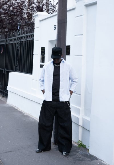 Camisa blanca de botones sobre una camiseta negra, combinada con pantalones de mezclilla anchos negros. Los accesorios incluyen una cadena de plata y un sombrero negro.