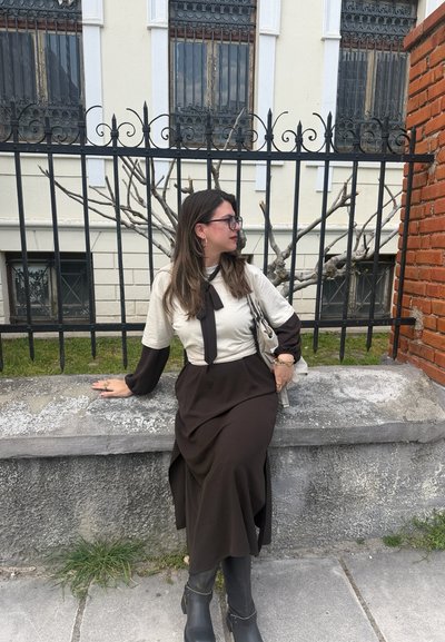 Mujer con un vestido largo en blanco y negro y gafas sentada en un borde de piedra frente a una valla de hierro negra y un árbol desnudo.