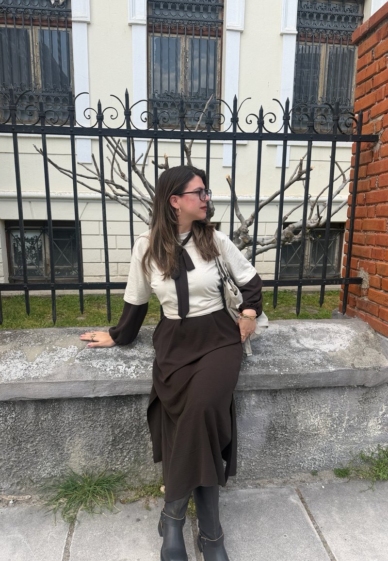 Ženska v dolgi črno-beli obleki z očali sedi na kamnitem robu pred črno železno ograjo in golim drevesom.