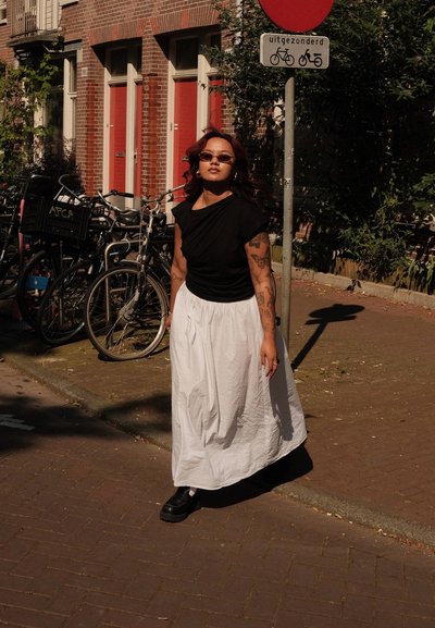 Top negro de manga corta combinado con una falda larga y fluida blanca hasta los tobillos. Zapatos de plataforma negros. El fondo presenta bicicletas y edificios de ladrillo.