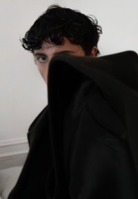 Schwarzer Stoff verbirgt eine Figur mit einer glatten, drapierten Textur. Weiches, welliges, dunkles Haar ist sichtbar, ebenso wie ein teilweise verborgenes Gesicht und ein Auge.