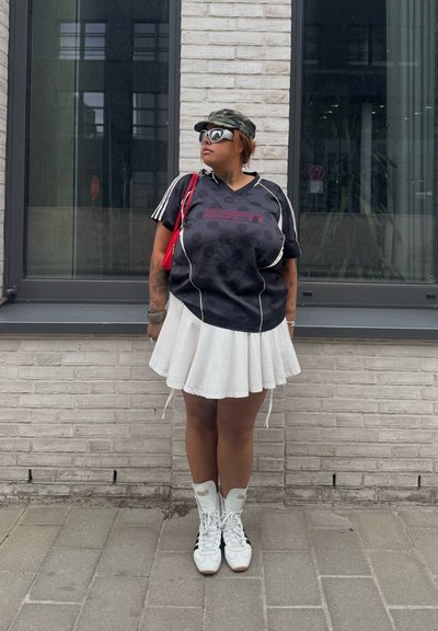 Persona con camiseta deportiva negra, falda plisada blanca, zapatillas blancas, gafas de sol y gorra de camuflaje, de pie frente a una pared de ladrillos con ventanas.