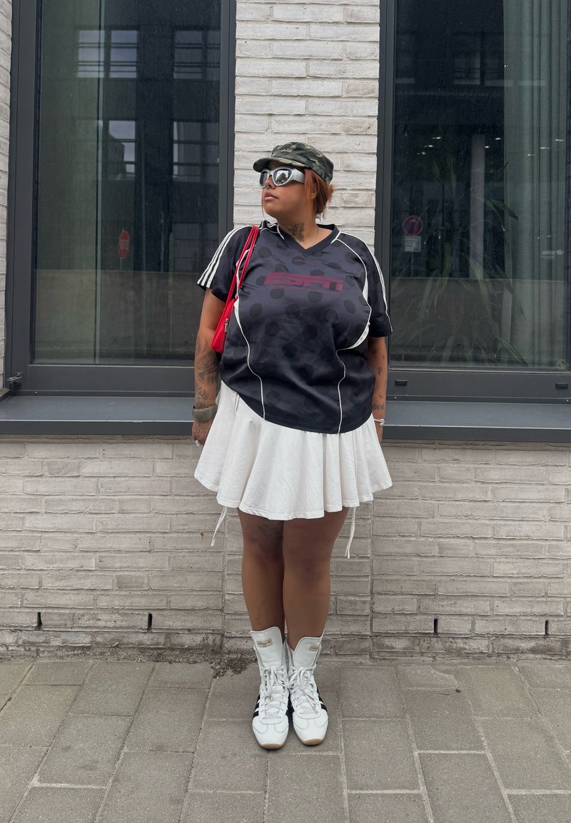 Person trägt ein schwarzes Sportshirt, weißen plissierten Rock, weiße Sneaker, Sonnenbrille und eine Camouflage-Kappe und steht vor einer Backsteinmauer mit Fenstern.