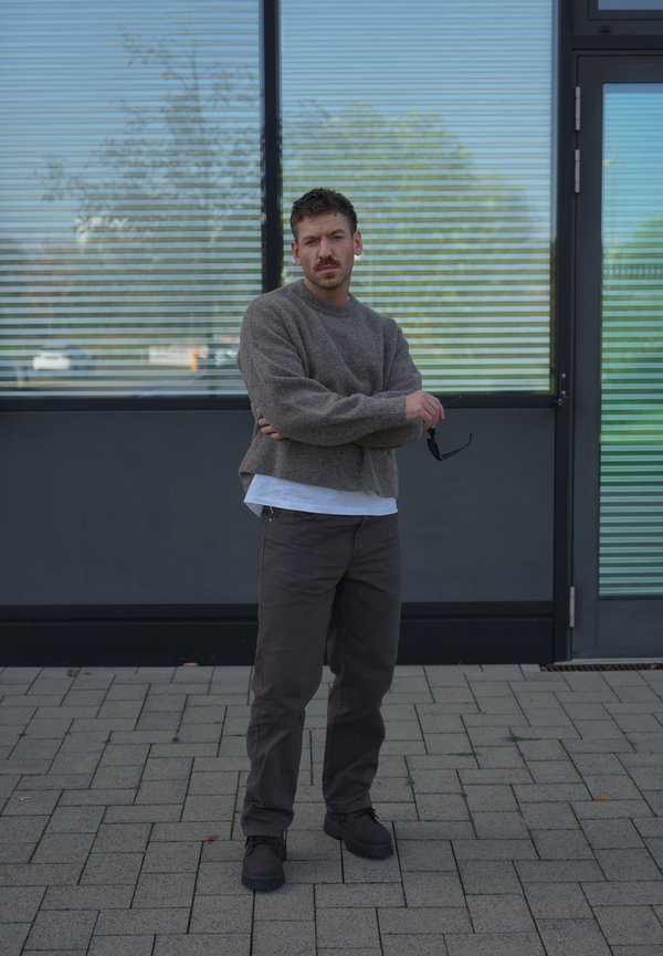 Un homme en pull gris et pantalon foncé se tient les bras croisés devant un bâtiment moderne, en tenant des lunettes de soleil.