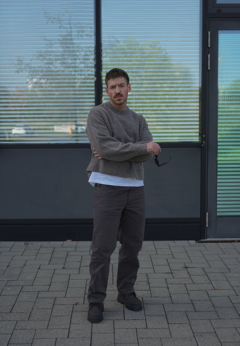 Un homme en pull gris et pantalon foncé se tient les bras croisés devant un bâtiment moderne, en tenant des lunettes de soleil.