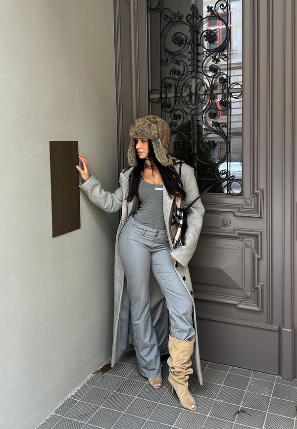 Femme portant un chapeau en fourrure, un long manteau, un haut gris et des bottes, appuyée contre un mur près d'une porte en métal ornée avec une plaque vierge.