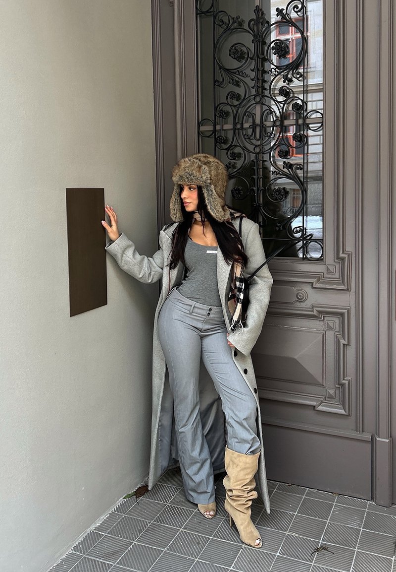 Femme portant un chapeau en fourrure, un long manteau, un haut gris et des bottes, appuyée contre un mur près d'une porte en métal ornée avec une plaque vierge.