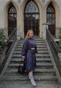 Felpa blu scuro oversize, gonna nera plissettata, sneakers bianche con calzini colorati, che tiene una borsa nera, in piedi su gradini di pietra vicino a un edificio.