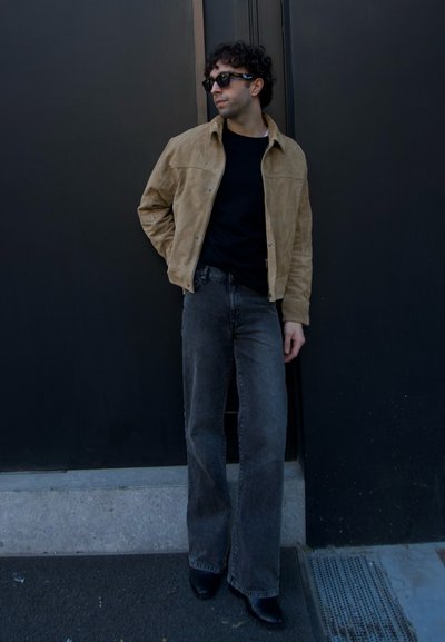 Chaqueta de ante marrón sobre una camiseta negra, combinada con jeans gris de cintura alta y botas negras. La persona lleva gafas de sol y está de pie contra una pared oscura.