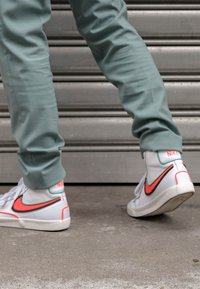 Vita högtoppsneakers med en rosa och grön Nike-logga, tillsammans med ljusgröna byxor, mot en texturerad grå bakgrund.