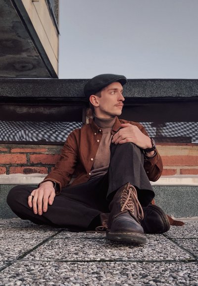 Camisa marrón de botones sobre un cuello de tortuga beige, pantalones negros y botas de cuero marrón con cordones. Sentado sobre una superficie texturada con fondo de ladrillo.