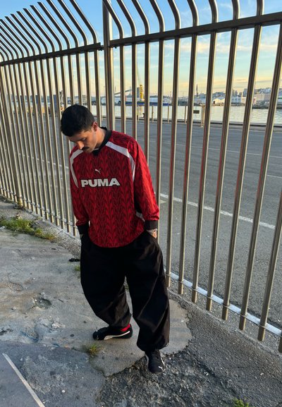 Jersey de manga larga de Puma con patrón rojo y acentos blancos, combinado con pantalones negros holgados y zapatillas negras. Escenario junto a una valla de metal.
