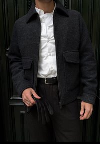 Giacca di lana grigio scuro con due tasche frontali, chiusura con cerniera e colletto. Abbinata a una camicia bianca e con un guanto di pelle nero in mano.