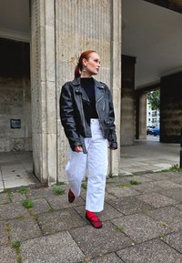 Must nähkida nahkvelk mustade turtleneckiga, valged laiad püksid ja punased kingad musta võrgu detailiga. Tekstuurse kivi taust.