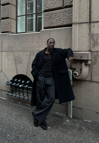 Hombre negro con gafas redondas, abrigo negro y pantalones anchos apoyado contra una pared de piedra texturizada cerca de una ventana con rejas en una calle de la ciudad.