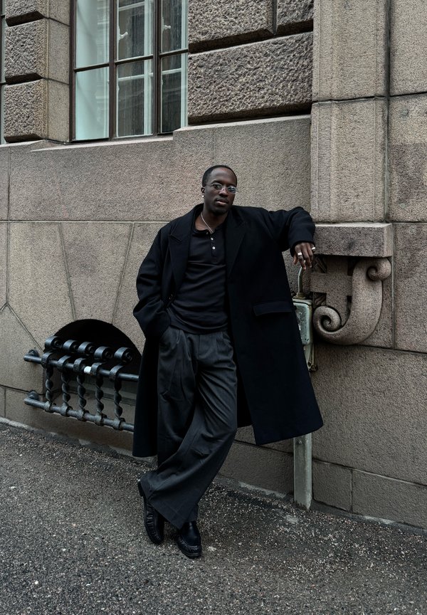 Musta mies pyöreät silmälasit, musta takki ja leveälahkeiset housut nojaa karkeapintaiseen kiviseinään aidatun ikkunan lähellä kaupungin kadulla.