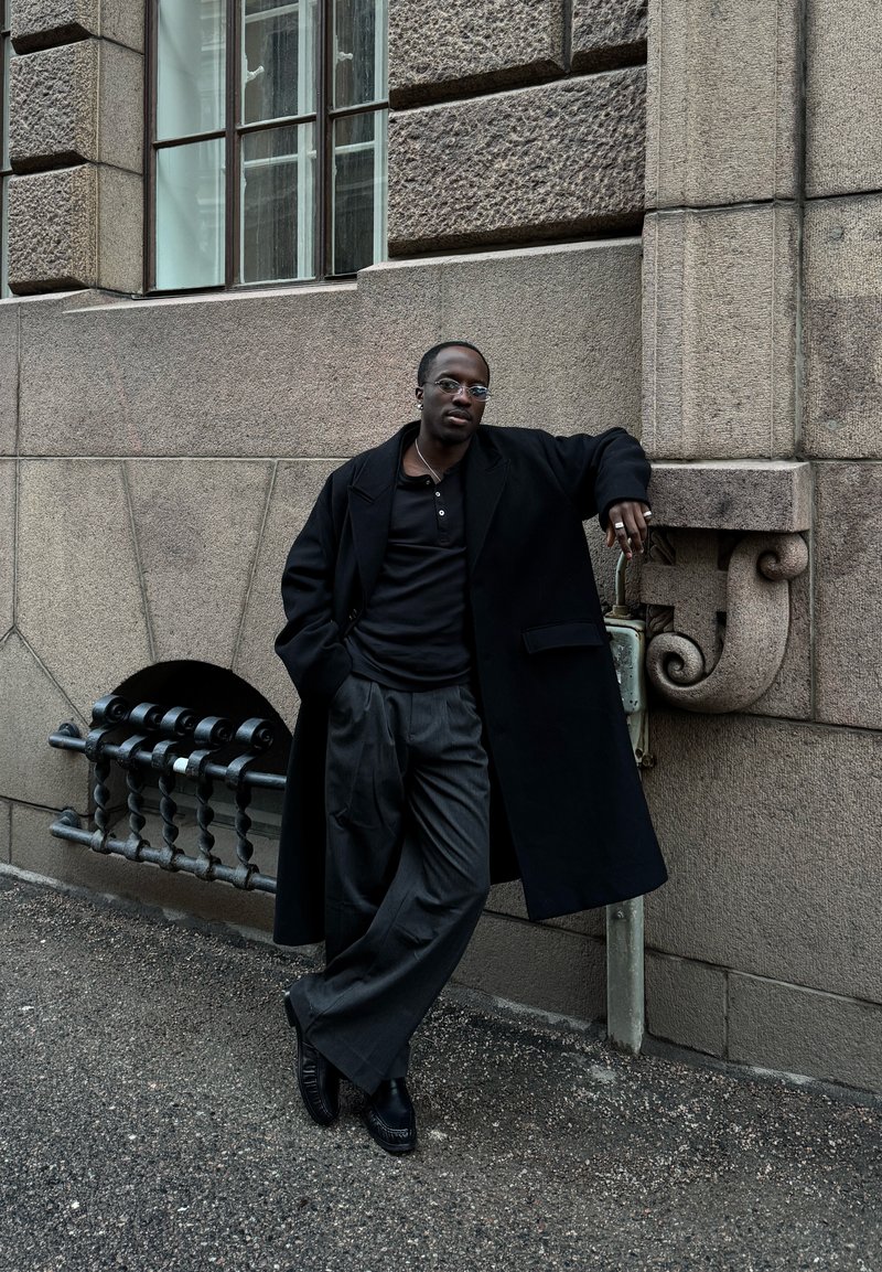 Schwarzer Mann mit runden Brillen, schwarzem Mantel und weitem Hosen lehnt an einer strukturierten Steinmauer in der Nähe eines vergitterten Fensters in einer Straßenszene.