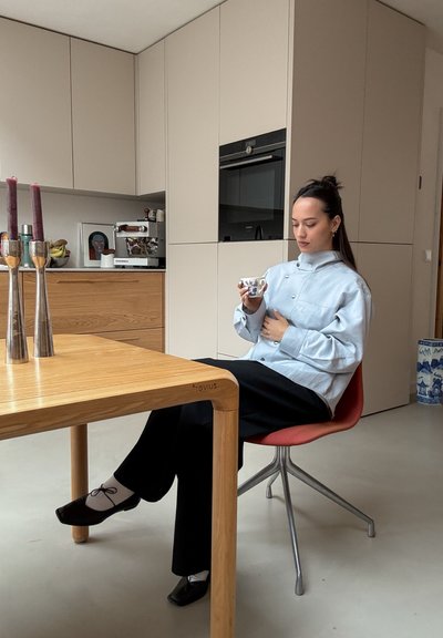 Camisa azul claro de gran tamaño, pantalones negros, sentado en una silla roja junto a una mesa de madera. Sosteniendo una taza con estampado azul. Candelabros plateados cerca.