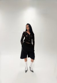 Zwarte mesh top met lange mouwen en knopen, gecombineerd met losse zwarte shorts met witte strepen, witte enkelsokken en zwarte schoenen met hak.