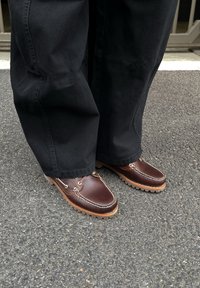 Hnědé kožené boty s kulatou špičkou, kontrastním prošitím a gumovou podešví. Boty mají tkaničky a jsou nošeny s černými kalhotami.