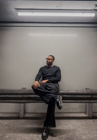 Haut noir à manches longues avec détail à boutons, pantalon large noir, et chaussures noires ; assise sur un banc dans un cadre intérieur minimaliste.