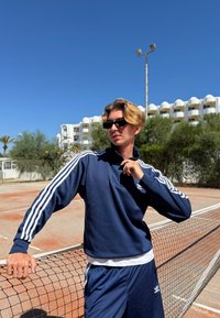 Felpa sportiva blu navy con zip e strisce bianche sulle maniche, abbinata a pantaloni corti coordinati. Indossata con occhiali da sole neri su un campo da tennis illuminato dal sole.