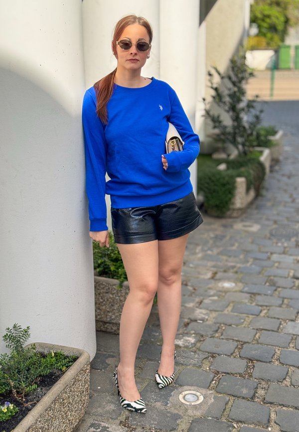 Žena u plavoj majici dugih rukava, crnim kožnim kratkim hlačama, štiklama sa uzorkom zebre i sunčanim naočalama, naslanja se na bijeli stup na kaldrmisanoj stazi.