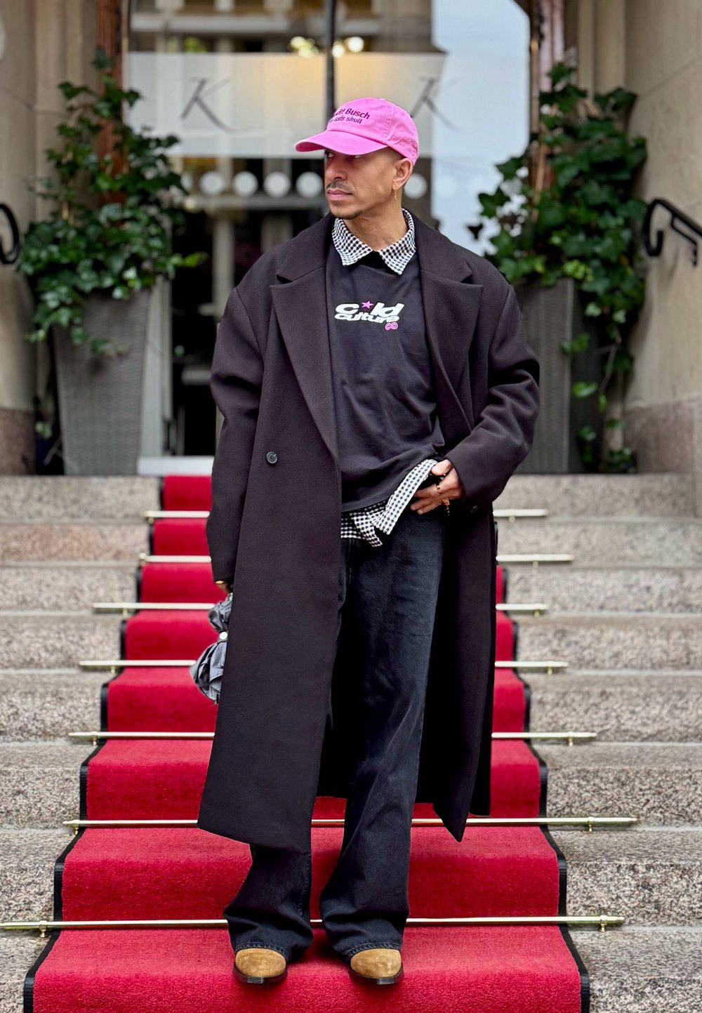 Melnas garas mēteles pār melnu grafisku kapuci, četrkantaina krekla maliņas redzamas, melnas bikses un gaiši brūnas kurpes uz sarkanu paklāja kāpnēm. Valkāts rozā cepure.