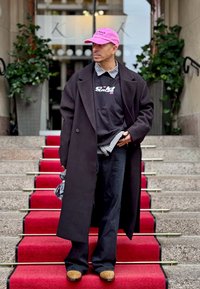 Melnas garas mēteles pār melnu grafisku kapuci, četrkantaina krekla maliņas redzamas, melnas bikses un gaiši brūnas kurpes uz sarkanu paklāja kāpnēm. Valkāts rozā cepure.