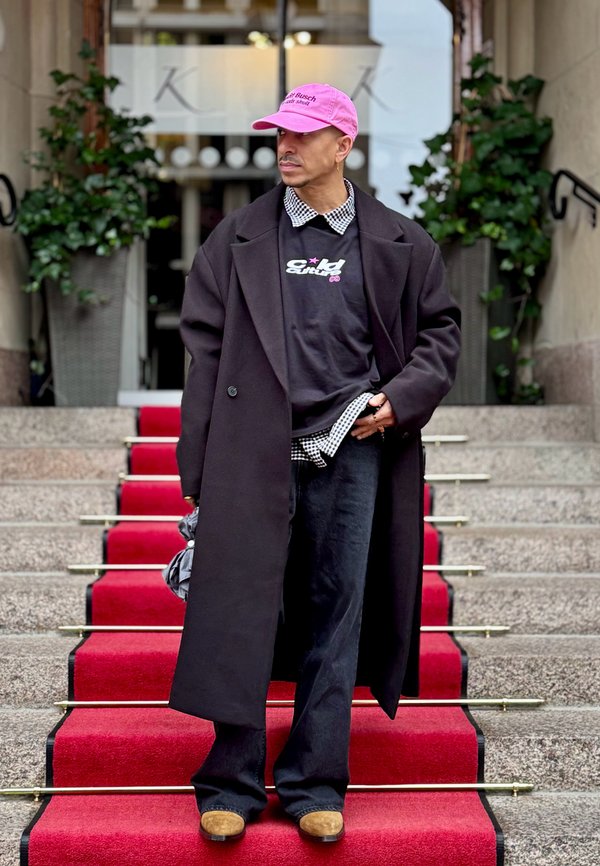 Pardesi lung negru over un hanorac grafic negru, o cămașă cu carouri care se vede, pantaloni negri și pantofi călbui pe niște scări tapisate cu covor roșu. Poartă o șapcă roz.