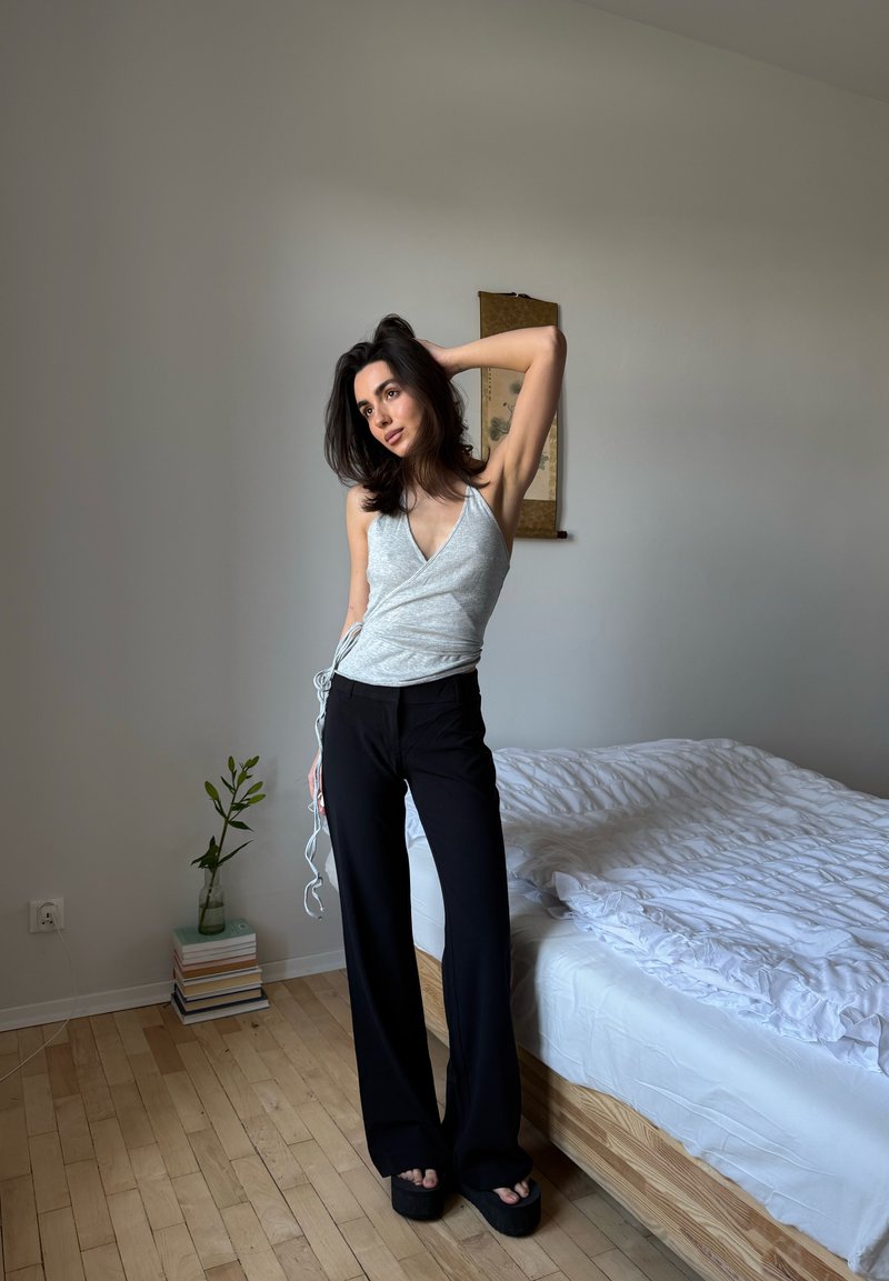 Femme portant un haut gris sans manches et un pantalon noir, posant avec une main dans les cheveux à côté d'un lit dans une pièce décorée de manière minimaliste.