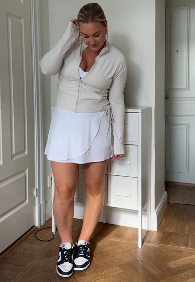 Chaqueta de chándal beige claro con cremallera, de manga larga y diseño ajustado, combinada con una falda envolvente blanca y zapatillas en blanco y negro.