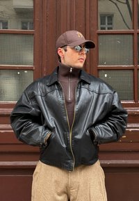 Jeune homme portant une casquette brune, de petites lunettes de soleil, un pull zippé marron, une veste en cuir noire et un pantalon beige, debout les mains dans les poches.