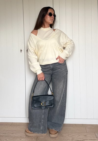 Suéter crema de gran tamaño con aberturas en los hombros, combinado con jeans de pierna ancha gris y un bolso negro. Zapatos de plataforma marrones completan el look.