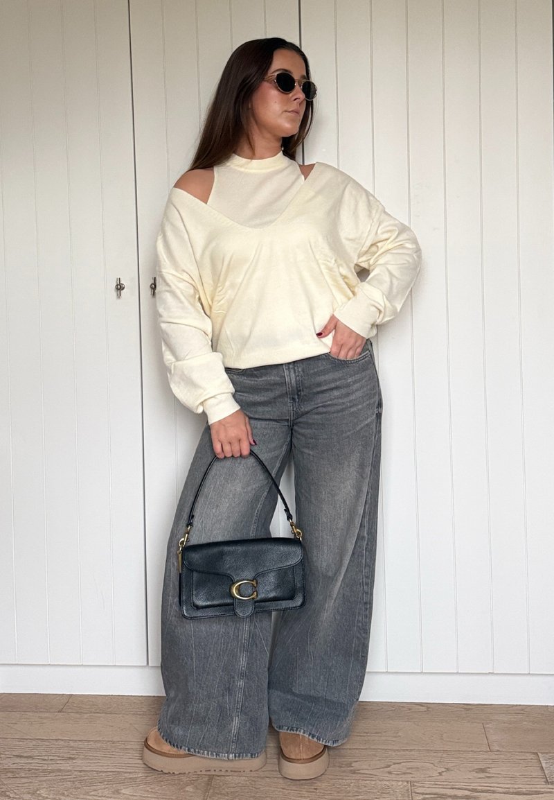 Camisola creme oversized com recortes nos ombros, combinada com jeans cinzentos de perna larga e uma mala preta. Sapatos plataforma castanhos completam o visual.