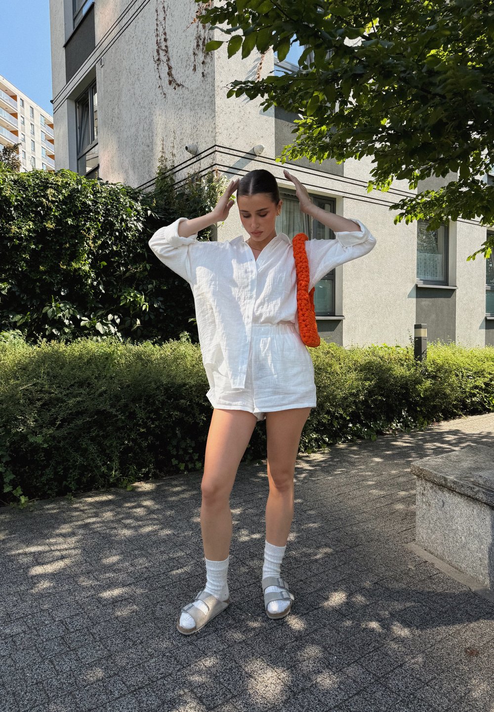 Λευκό πουκάμισο από λινό με κουμπιά και σορτς, πορτοκαλί υφασμάτινη τσάντα, ασημένια σανδάλια και λευκές κάλτσες; εξωτερικό περιβάλλον με πράσινο και κτίρια.