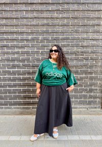 Persona con cabello largo y rizado, llevando gafas de sol negras, camiseta verde de Adidas, falda larga negra y zapatillas blancas, de pie junto a una pared de ladrillos grises.